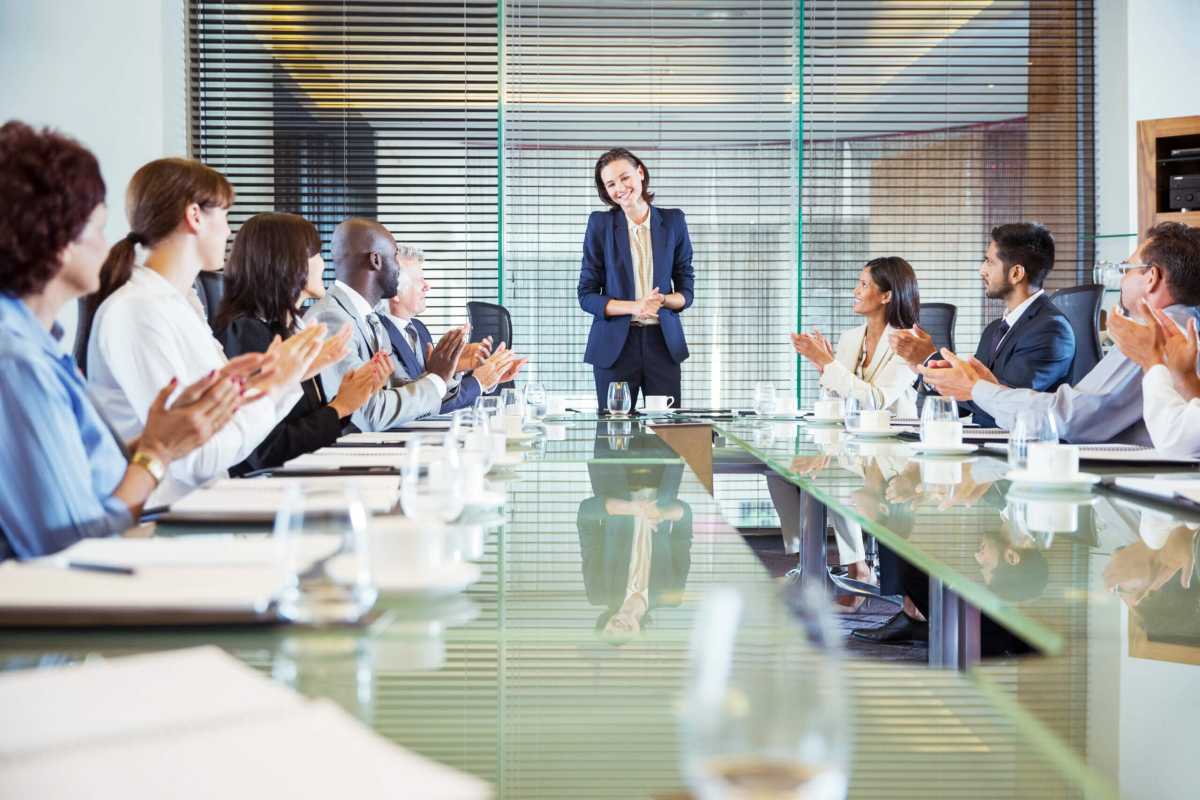 Conference participants applauding to young businesswoman standing at head of conference table