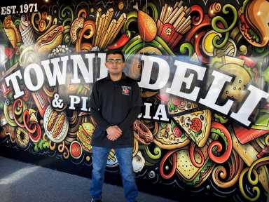 man in a black shirt standing in front of Towne Deli & Pizzeria sign on Staten Island