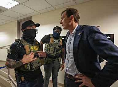 Man in mask looks back at man in suit, with latter providing a stern gaze and hands on hips