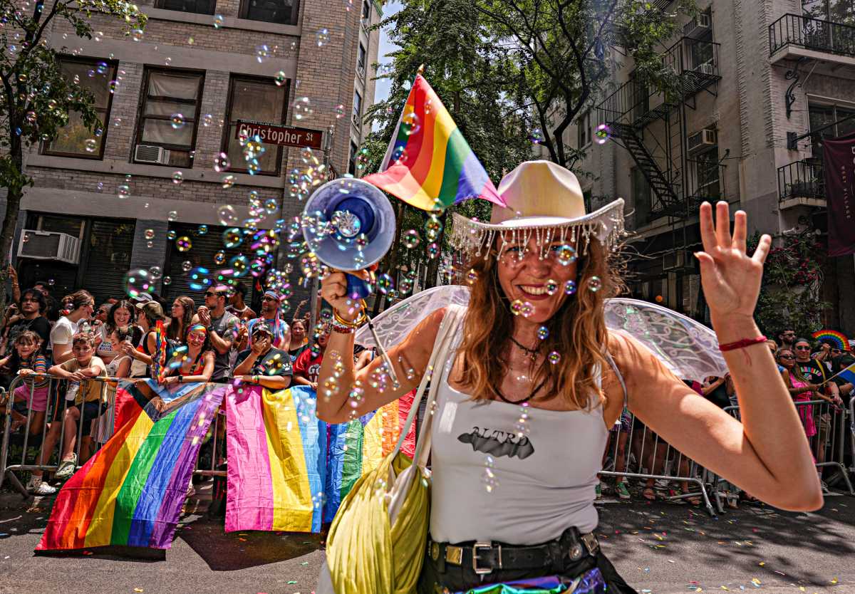 Woman in tanktop and cowboy hat using bubble gun with rainbow Pride flag waving in the background.