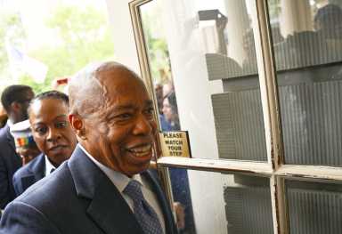 Mayor Eric Adams smiling and wearing suit as he walks near glass door