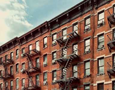 What would a rent freeze mean for NYC if mayoral candidates and advocates get their way? 15 This image showcases a timeless urban scene of a brick apartment building adorned with iconic fire escapes. The warm tones of the brick contrast beautifully with the soft blue sky, capturing the essence of city life in a historical neighborhood. The architectural details, including the ornate cornices and window air conditioning units, evoke a sense of nostalgia and urban charm. Perfect for projects related to city living, historical architecture, or urban photography