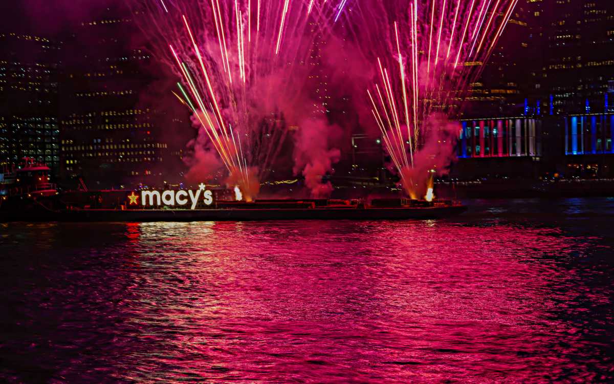Red fireworks set off from a barge in water with the Macy's logo
