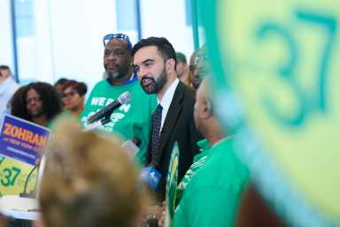 man in beard and suit speaks in microphone with green union sign with 37 in foreground at Mamdani rally