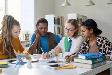 Young female teacher sitting at table with multiethnic group of students during class in school or college