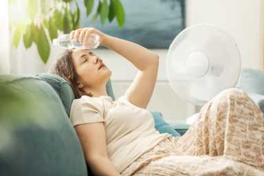 Young woman sitting on the couch at home and keeping a fresh water bottle on her forehead, she is suffering from the heat