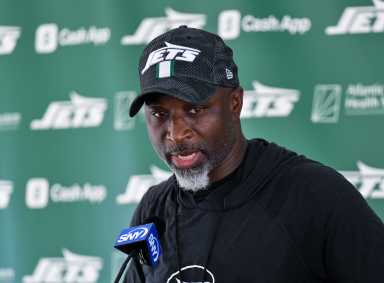 man in Jets cap and shirt against green backdrop speaks into microphone