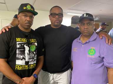 three men standing in a room, two wearing black shirts, one wearing a purple shirt