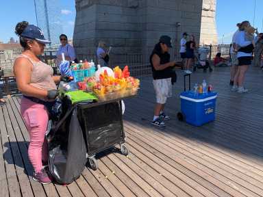 Defying a NYC ban, illegal vending thrives on iconic Brooklyn Bridge this summer 9 people selling items on a bridge with a wooden floor